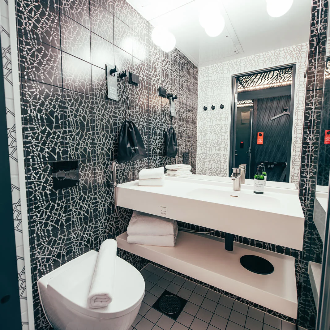 A modern bathroom features a white sink with a faucet, white towels, and an adjacent toilet. The walls have a black geometric pattern, and a mirror reflects more surroundings.