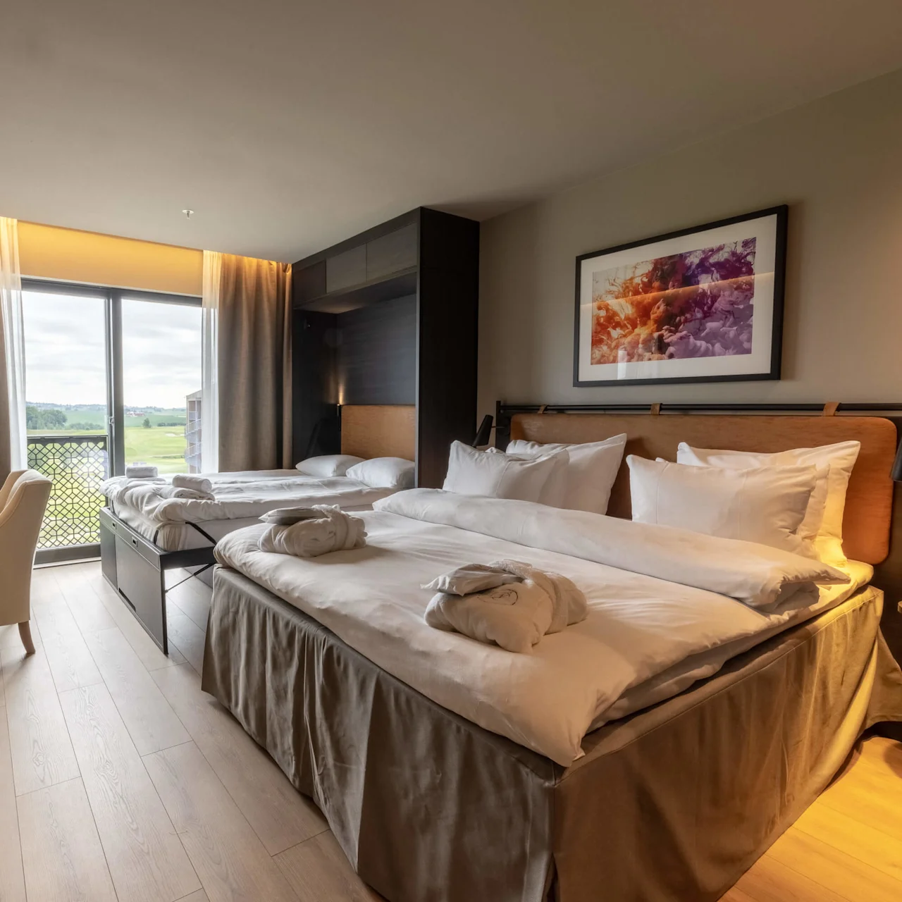 Ett välupplyst modernt hotellrum med två sängar, konst på väggen och utsikt över grönska utanför fönstret.