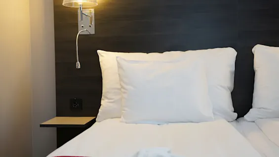 Mukava sänky valkoisilla liinavaatteilla ja lukuvalolla huoneessa Quality Hotel Sundsvallissa, valmiina rentouttavaan oleskeluun.