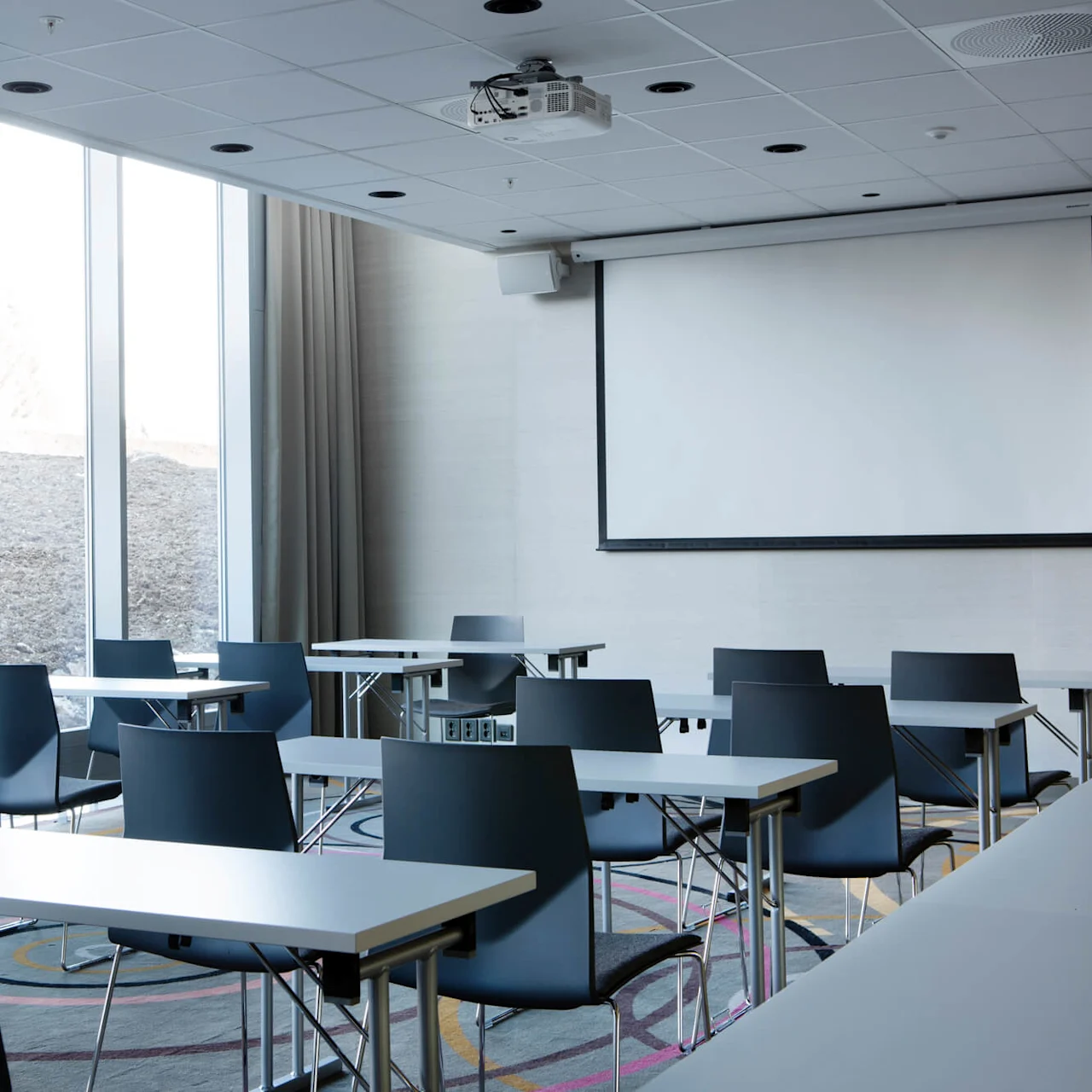Classroom with empty desks and chairs, featuring a large projection screen. Sunlight streams through tall windows, with beige curtains partially open, brightening the carpeted room.