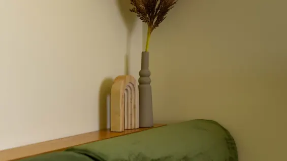 En grön kudde vilar på en träbänk bredvid en hög vas med torkat gräs, mot en ljus vägg med en inramad abstrakt konstverk ovanför.