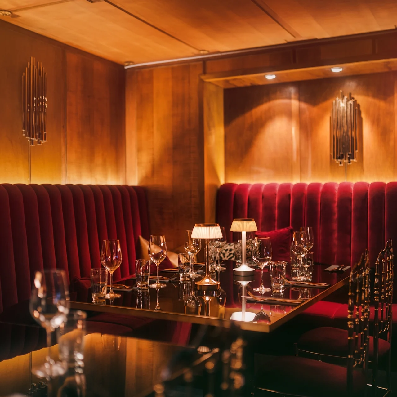 A dimly lit restaurant booth with red velvet seating and a set dining table featuring neatly arranged glasses, cutlery, and small lamps, surrounded by warm wooden paneling.