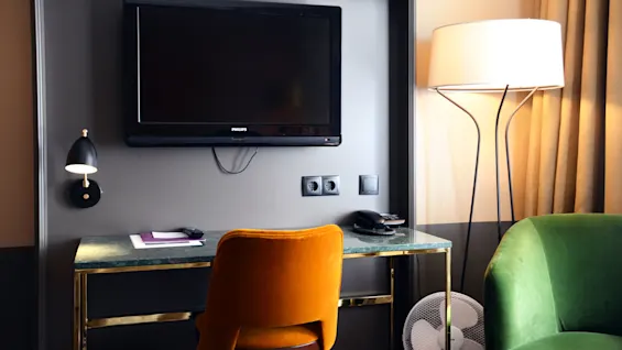 A mounted TV above a desk with a chair, surrounded by a wall lamp, phone, and a standing lamp in a room.