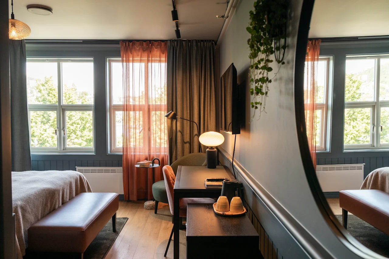 Ett mysigt hotellrum har en säng och ett skrivbord; stora fönster med tunna gardiner släpper in naturligt ljus. En spegel reflekterar en del av rummets inredning och utsikt.