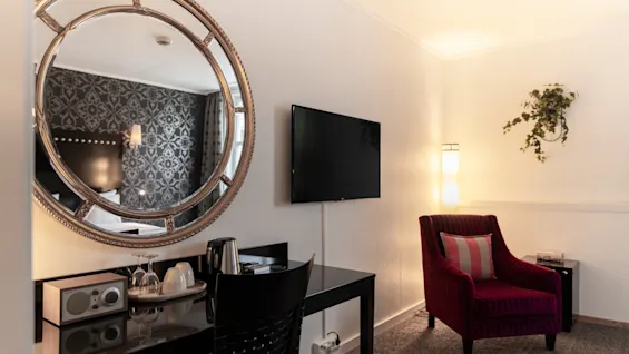 Viihtyisä hotellihuone, jossa on työpöytä peilillä, seinälle asennettu televisio ja mukava nojatuoli tyynyllä.