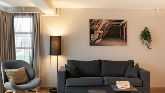 En koselig stue med en mørk sofa, lenestol, gulvlampe og et bilde av trebjelker på veggen.