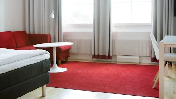 A red sofa sits next to a white round table in a bright room with grey curtains, a radiator, and a neat bed.