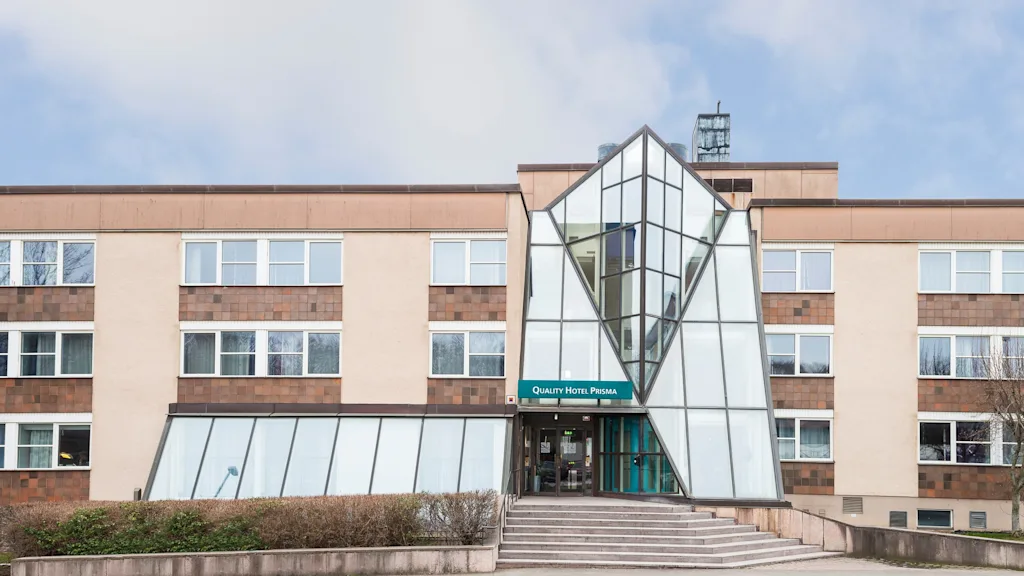 En modern hotellentré med glasfasad och geometrisk design, märkt "Quality Hotel Prisma," omgiven av buskar och ett stenbelagt område under en delvis molnig himmel.