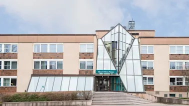 En modern hotellentré med glasfasad och geometrisk design, märkt "Quality Hotel Prisma," omgiven av buskar och ett stenbelagt område under en delvis molnig himmel.