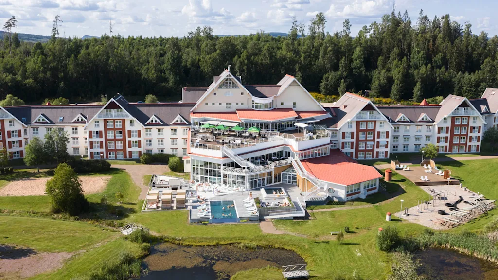 A large hotel with red roofs and white walls has guests enjoying an outdoor pool and sunbathing. Surrounded by lush green forest, it includes a patio with seating.