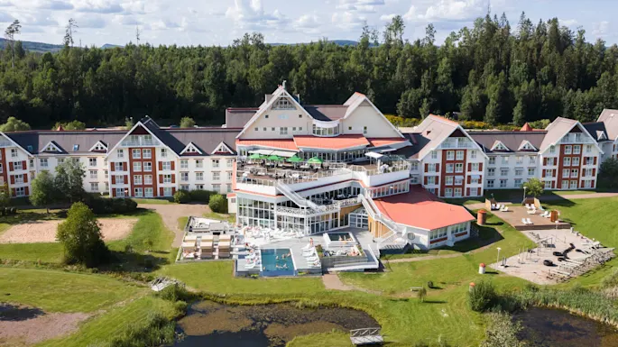 Et stort hotell med røde tak og hvite vegger har gjester som nyter et utendørs svømmebasseng og solbading. Omgitt av frodig grønn skog med en terrasse med sitteplasser.