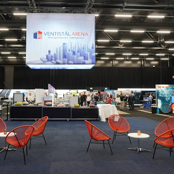 Orange chairs surrounding small tables are arranged on a carpeted floor, with people interacting at exhibition booths in a large, well-lit conference hall. Text: "VENTISTÅL ARENA Mulighetenes møteplass." 