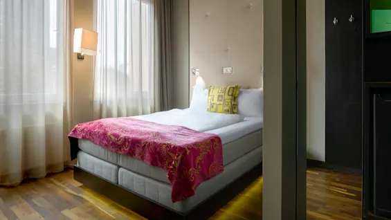 En pent oppredd seng med et fargerikt sengeteppe og mønstrede puter i et ryddig, godt opplyst hotellrom med gjennomsiktige gardiner og moderne dekor.