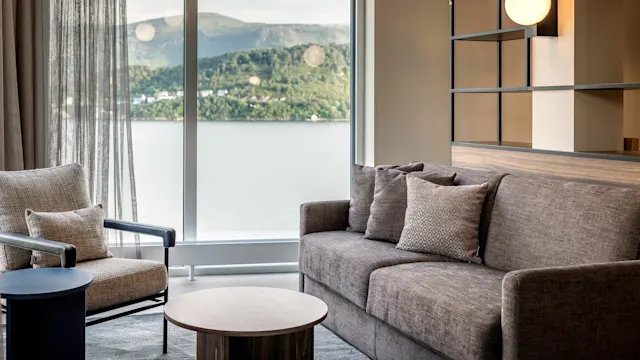 Brown sofa sits in a modern living room beside a chair and circular table, overlooking a scenic lake and mountain view through large windows.