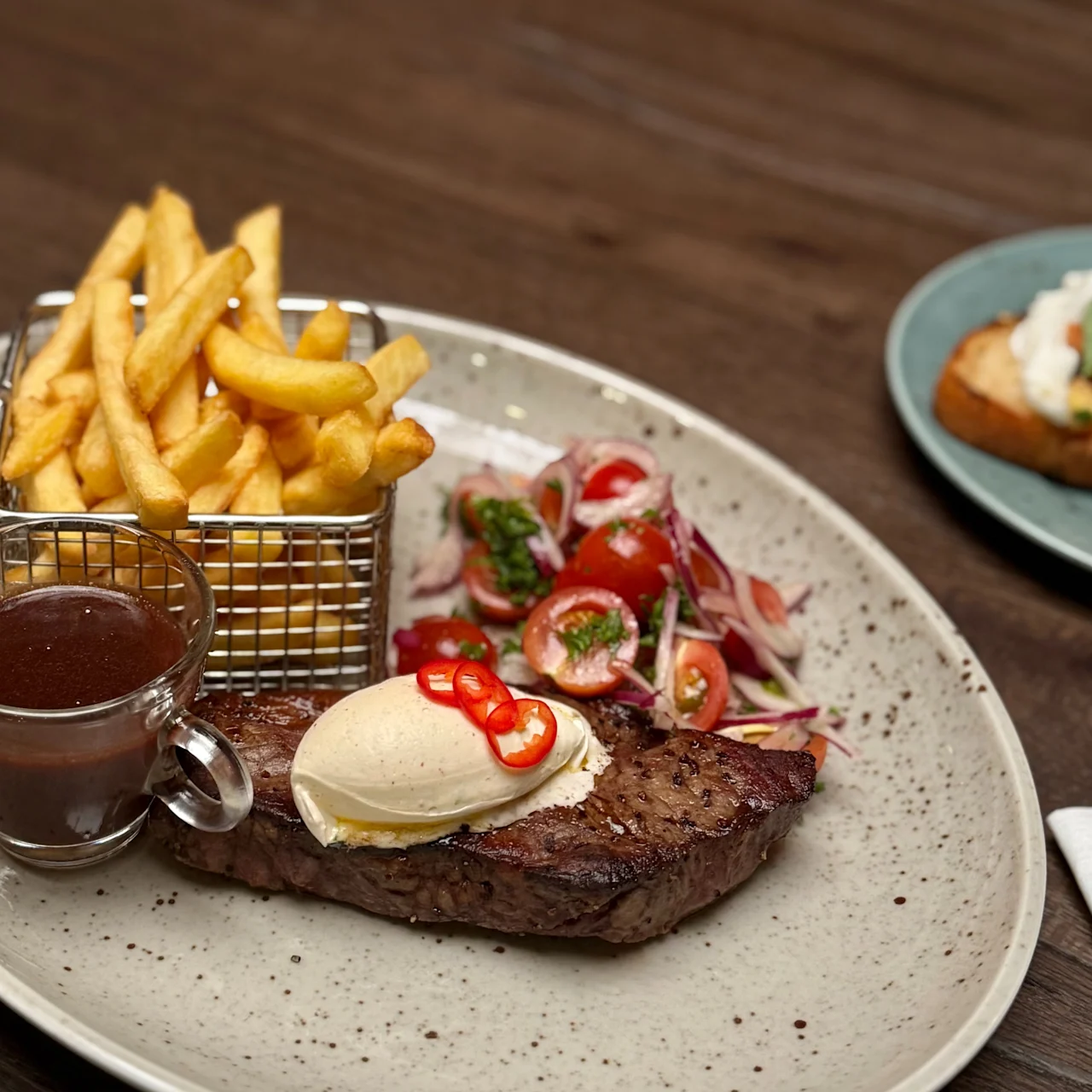 Steak topped with butter and chili peppers sits on a plate with a side salad and fries in a basket, accompanied by a small cup of sauce on a wooden table.