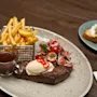Steak topped with butter and chili peppers sits on a plate with a side salad and fries in a basket, accompanied by a small cup of sauce on a wooden table.