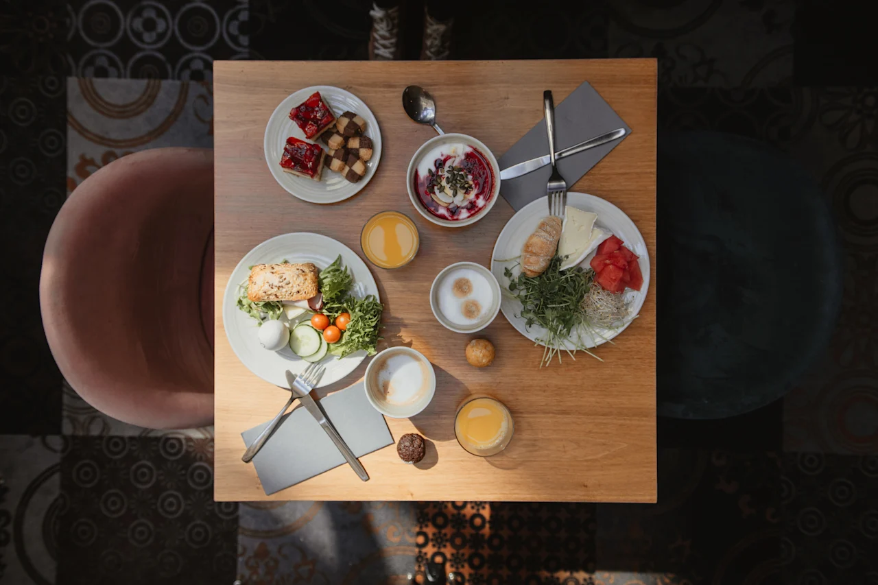 Frukosträtter ligger på ett träbord och visar bakverk, frukt och sallader med apelsinjuice och kaffe. En rosa stol och ett mörkt mönstrat golv omger upplevelsen.