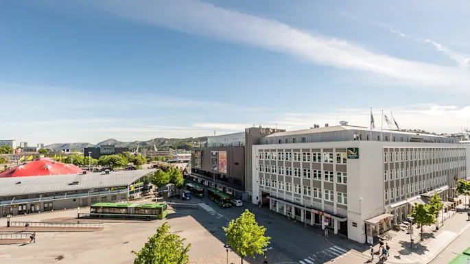 A large hotel building with a "Quality" sign stands beside a bus terminal with green buses. The scene is set in a sunny, urban environment with mountains in the background.