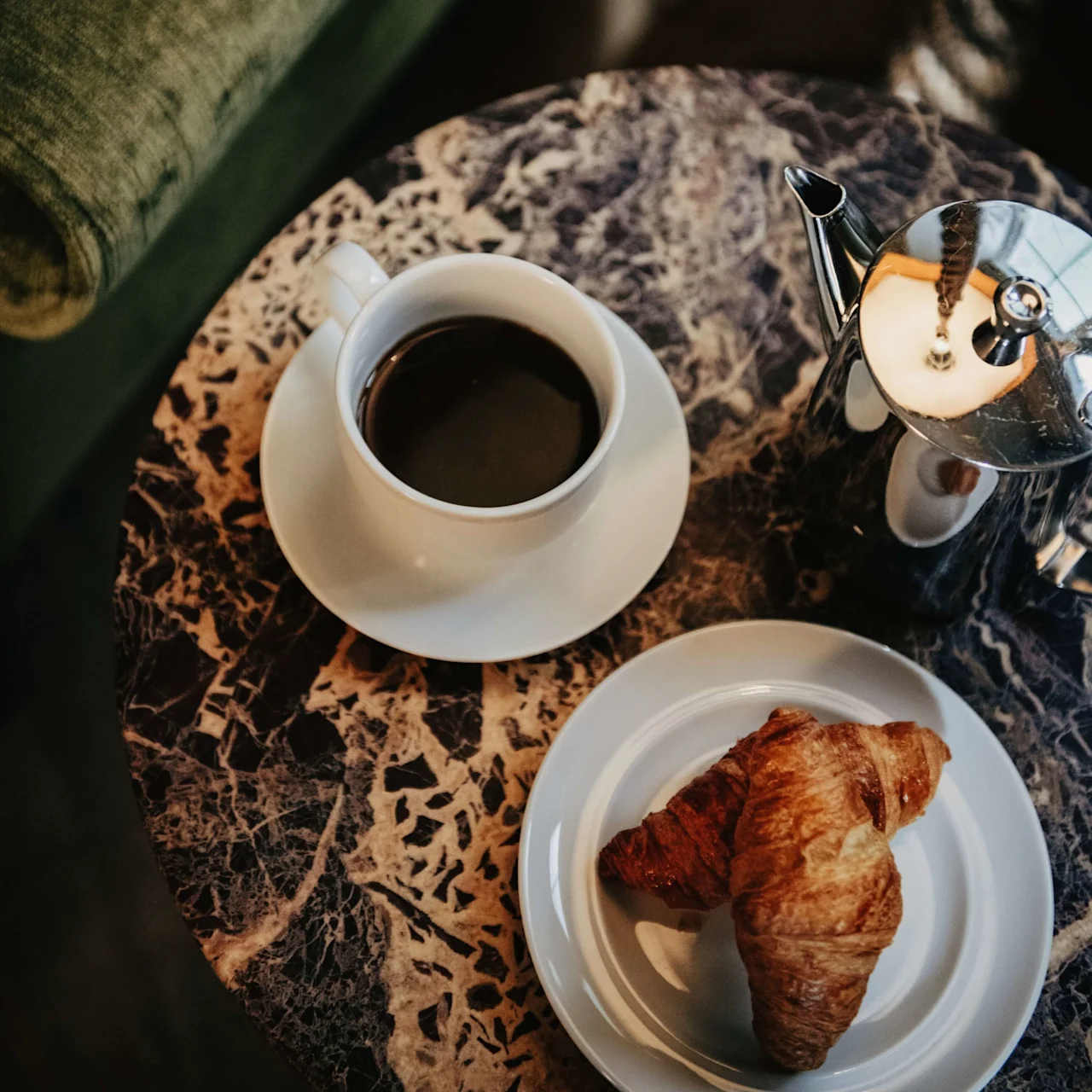 A cup of coffee, a croissant on a plate, and a silver coffeepot resting on a marbled table beside a green couch.