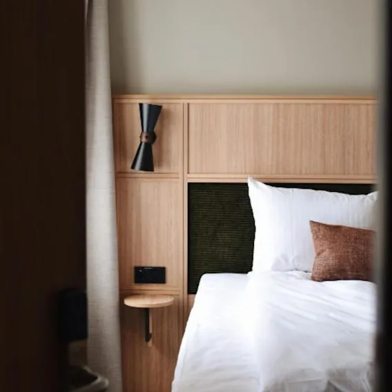 Glimt av et moderne hotellrom med oppredd seng i hvitt sengetøy og en lys sengegavl i tre.