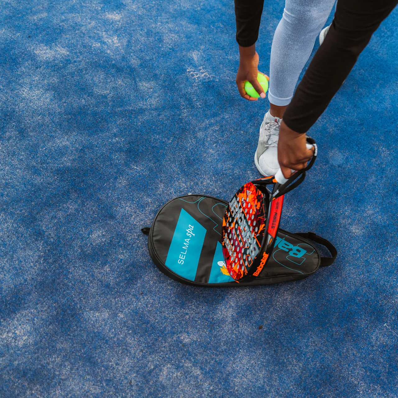 En tennisracket og ball holdt av en person som står på en blå overflate, nær en sportsbag merket "SELMA spa."