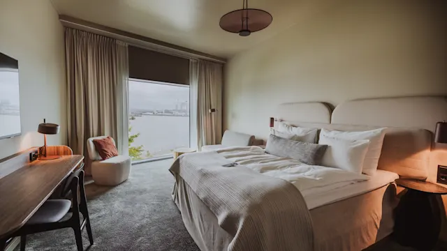 En pænt redt seng med hvidt sengetøj står i et moderne hotelværelse, flankeret af sideborde med lamper. Et stort vindue viser en naturskøn udsigt over vandet udenfor.