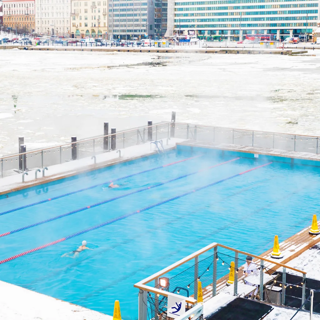 Helsinki in Winter, Allas Sea Pool. People swimming in a steaming outdoor pool surrounded by snow and ice with city buildings in the background.
