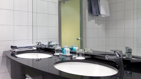 A modern bathroom vanity with two sinks, toiletries, and towels in a tiled room with a frosted glass door.
