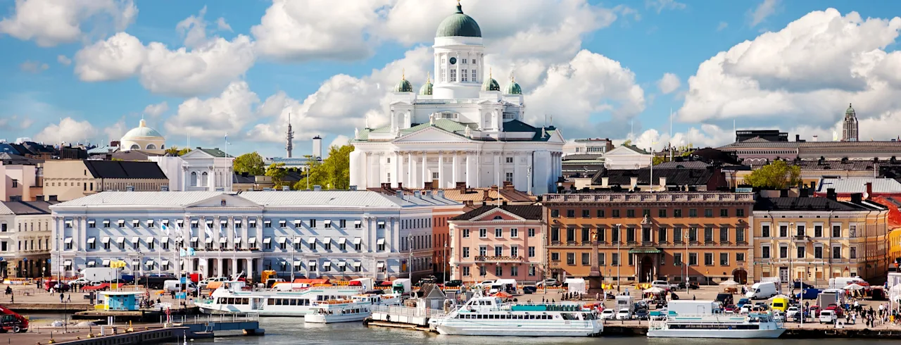 Båtar navigerar hamnen framför färgglada historiska byggnader och en stor vit katedral under en blå himmel med spridda moln.