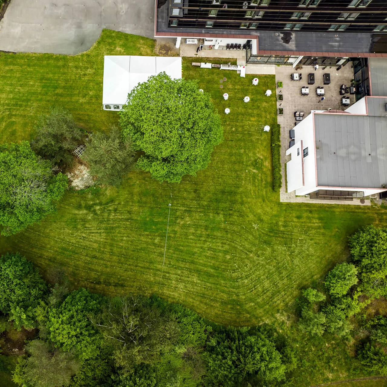 Aerial view of a grassy field with scattered trees and a white tent, adjacent to a building's patio with outdoor seating. Surrounded by dense foliage and a path.