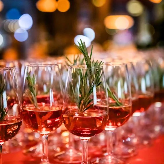 Rader av glas med rosa dryck och rosmarinkvistar på ett rött bord med suddiga ljus.