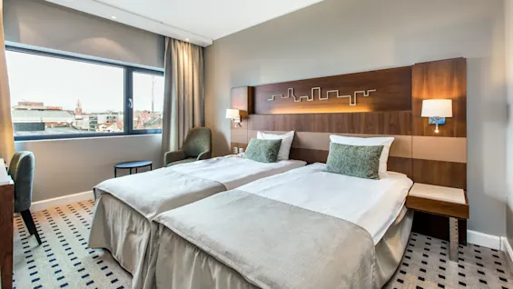 Two neatly made twin beds are positioned in a modern hotel room with large window, green armchairs, and wooden headboard. A cityscape view is visible outside.