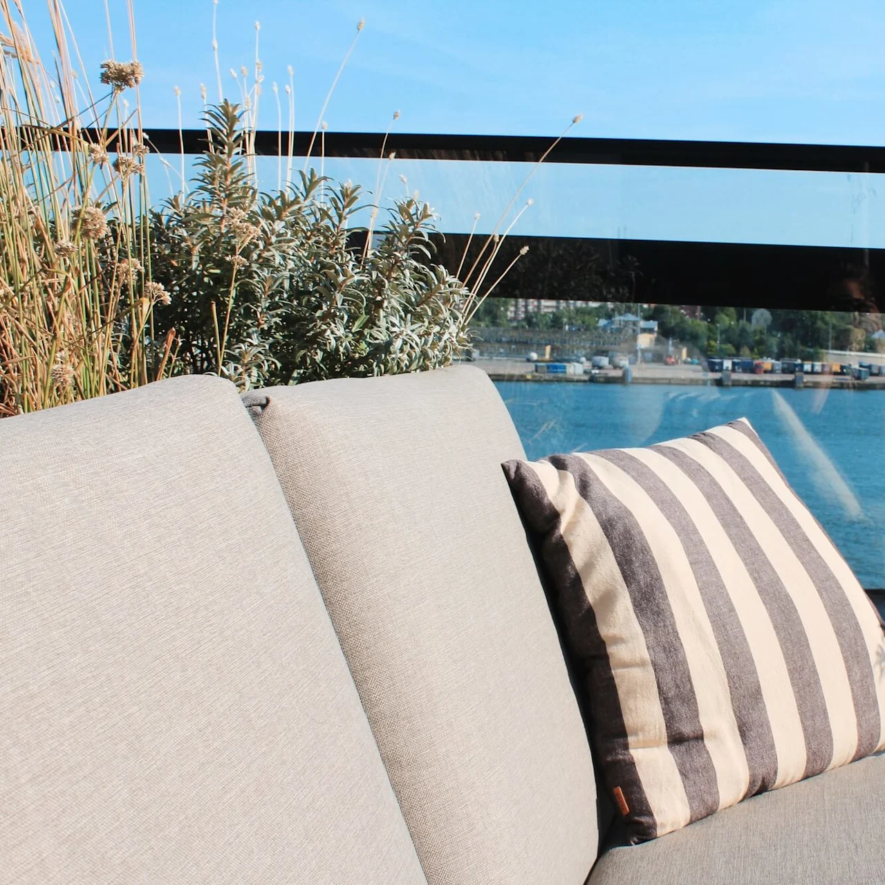A beige outdoor couch with a striped pillow is situated on a balcony overlooking a scenic waterfront, surrounded by potted plants and clear skies.