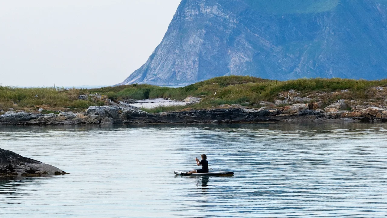 Kayaker paddling calmly across serene water, surrounded by rocky shoreline and distant mountainous landscape.