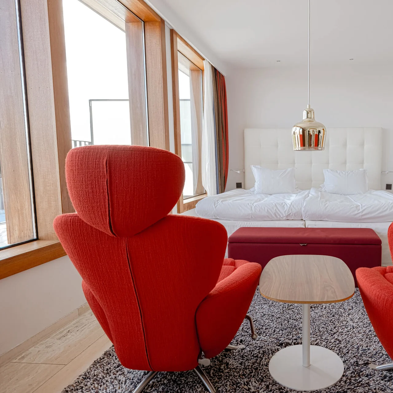 Two red chairs face a wooden table in a modern bedroom with large windows, white bedding, and a pendant light above, creating a cozy and stylish atmosphere.