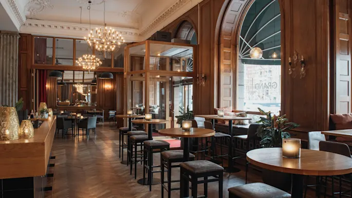 A warm and inviting hotel lobby bar with wooden interiors, comfortable seating, and large arched windows. GRAND is visible.