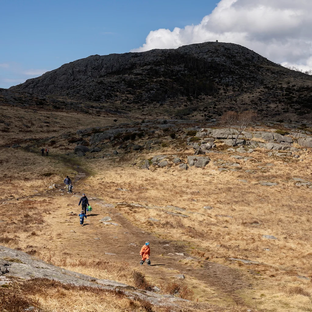 Perhe nauttii vaelluksesta mutkittelevalla hiekkatiellä kivisessä, ruohoisessa maisemassa suuren vuoren alla sinisen taivaan alla.