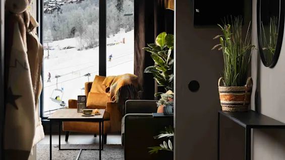 En hyggelig stue med en grøn sofa og en brun lænestol nær et vindue. Et snedækket landskab med skiløbere kan ses udenfor, og indendørs planter tilføjer grønt.