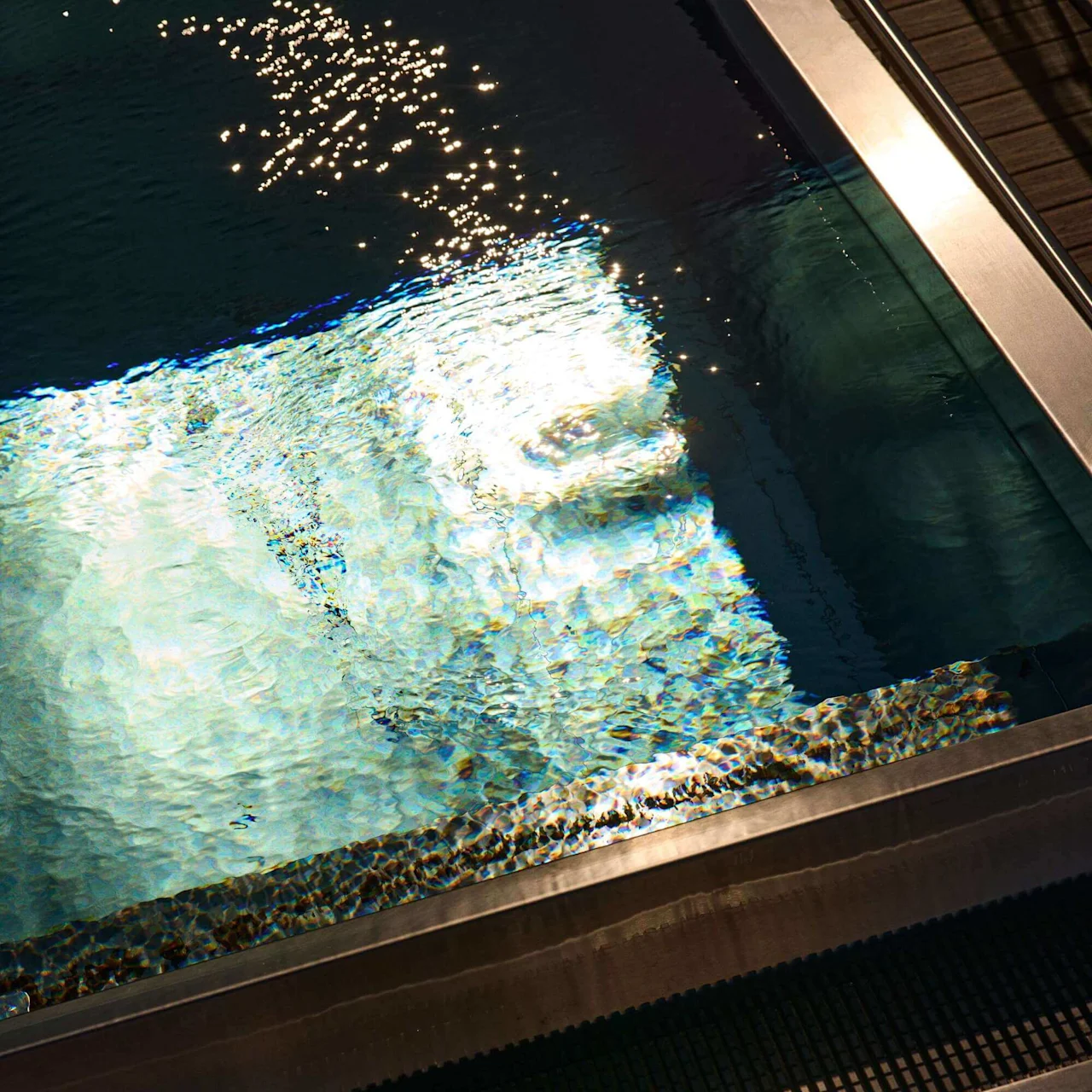 Pool water reflects light, creating shimmering patterns. The edge of the pool is bordered with metal and a black grate, suggesting an indoor swimming facility.
