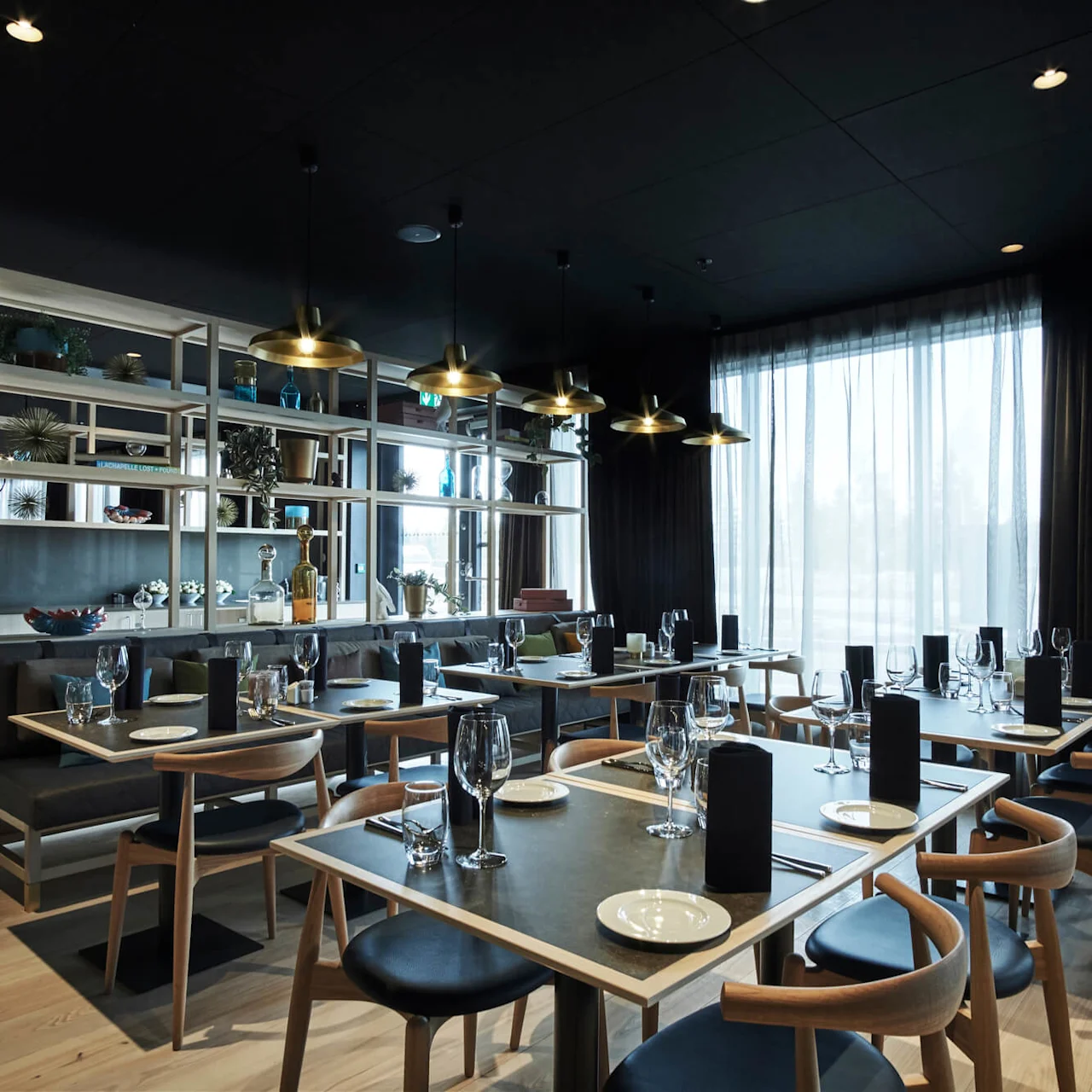 Modern dining tables set with black napkins and glassware, surrounded by wooden chairs in a sleek restaurant. Shelving with decorative items divides the space. Large windows let in natural light.