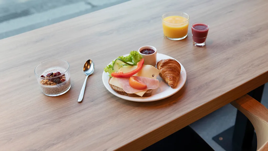 Puinen pöytä, jossa aamiaiskattaus sisältää chia-vanukasta, juusto- ja kasvisvoileipää, croissantin sekä kaksi lasillista appelsiini- ja marjamehua.