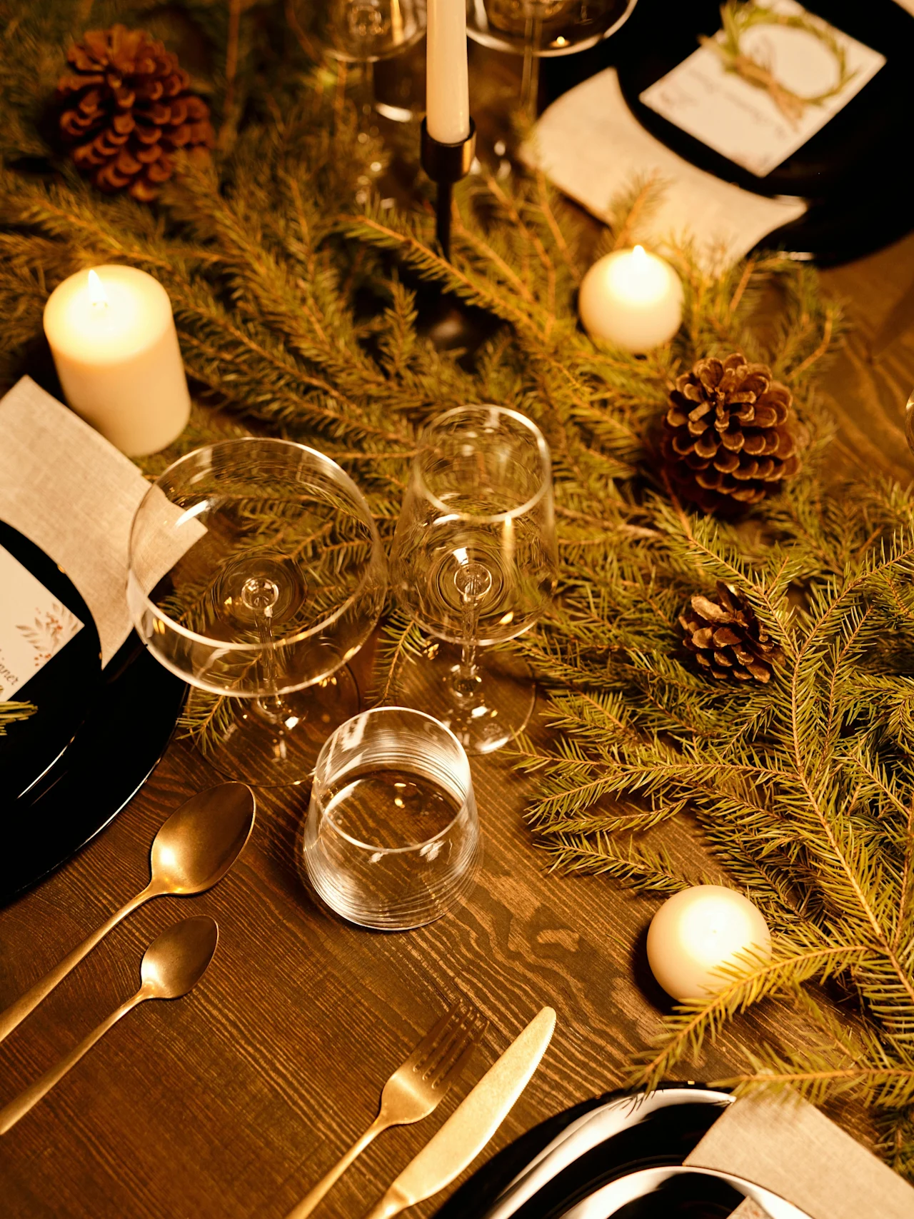 A dinner table is set with pine branches and pinecones, surrounded by candles and elegant cutlery, in a warm, festive ambiance. No text is visible.