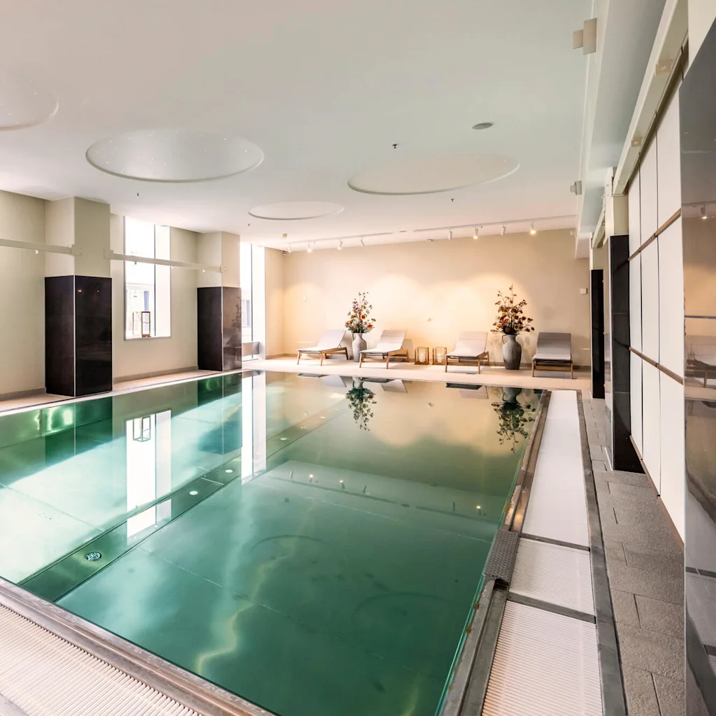 Swimming pool indoors, reflecting large windows on the left. Lounge chairs and decorative plants placed on the right side. Bright ceiling lights illuminate the modern, minimalist space.