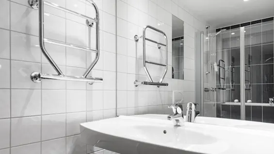 Sink with chrome faucet sits below towel racks attached to white tiled walls. A mirror reflects a shower area, enhancing the bright, modern bathroom atmosphere.