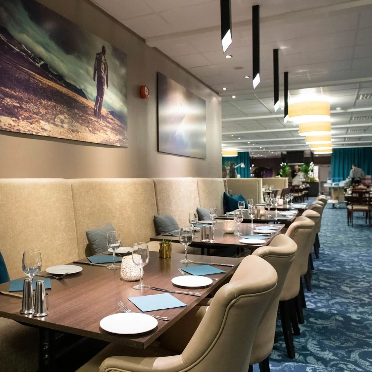 Ett långt matbord är dukat med vinglas och tallrikar på en vadderad bänk, omgiven av elegant inredning och konstverk i en rymlig, modern restaurang.
