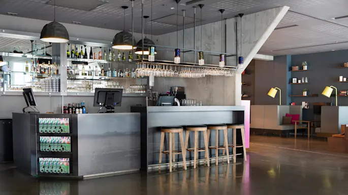 A modern bar area with wooden stools lined up at a sleek counter. Glasses hang overhead, and bottles are displayed behind. The surrounding area includes cozy seating with stylish lamps.