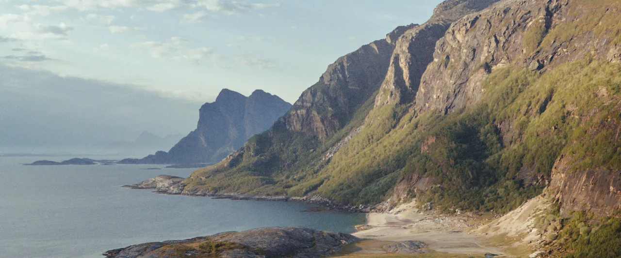 En båt flyter på klart turkost vatten nära en klippig strand med majestätiska berg i bakgrunden under en mjuk himmel.