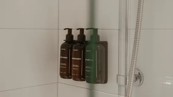 Flasker med pumper indeholder kropsvask, balsam og shampoo, monteret på en hvid flisebelagt brusevæg ved siden af en metalslange.