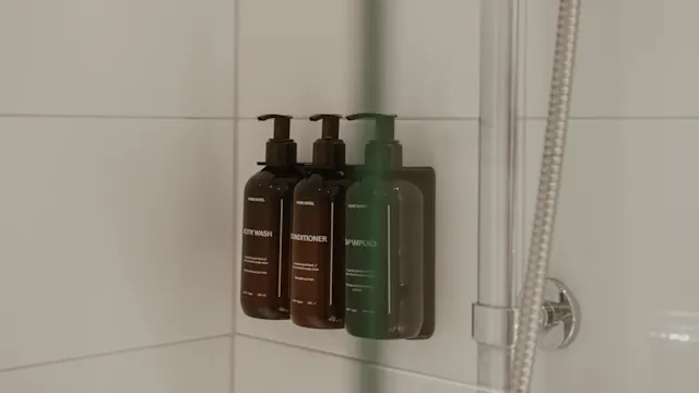 Flaskor med pumpar innehåller body wash, balsam och schampo, monterade på en vit kaklad duschvägg bredvid en metallslang.