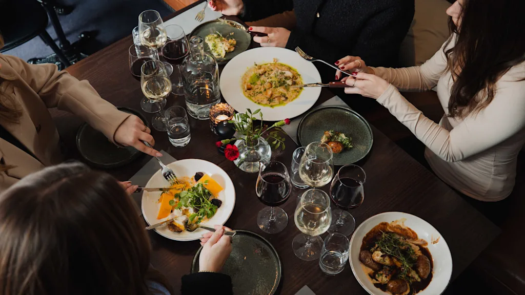 Fire personer spiser forskjellige retter med gafler i en restaurantsetting. Bordet har flere glass vin og vann blant dempet, koselig belysning.